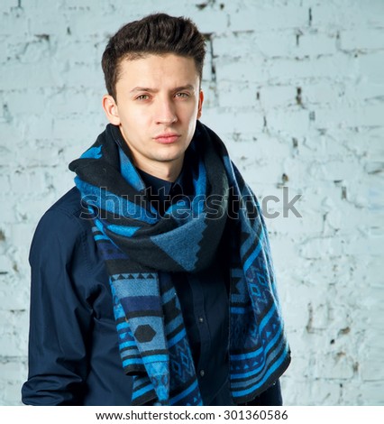 Portrait of a smart serious young man standing against blue background. Young serious man in a blue blouse and scarf. Autumn style of dress. A man in a warm scarf leaning on the wall of the room.