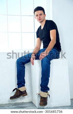 The man next to the window. One man sits with his back to the window. Fashionable male model. The empty space of the room around a man. Against the background of windows and light blue walls.