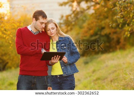 Couple in autumn garden. Man and woman together ordinary people. They are holding electronic tablet. Watch E-book. Emotional couple in love. Young couple using electronic tablet in autumn park.
