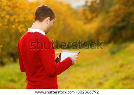 Young man using tablet computer on autumn street and city park. Man working on his tablet outdoors in the park. Modern man using tablet pc outside on a park bench.