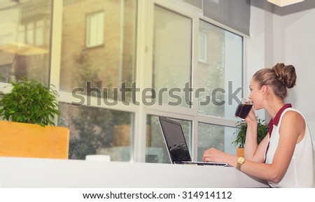 Woman with laptop. Young woman drinking morning coffee and works at a laptop in the office. Business woman - office worker. Girl student to work at a computer and drinking coffee. Look out the window.
