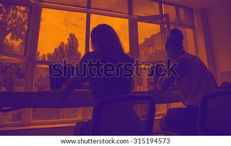 Silhouettes of business people on orange sunset. Silhouettes of two women working in front of the window. Two people communicating in dark office. Female student take an exam, or pass the test. Stress