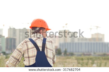 Man builder on the background of buildings under construction. A man in a construction orange helmet on his head standing with her back to the camera. A man in a half-turn.