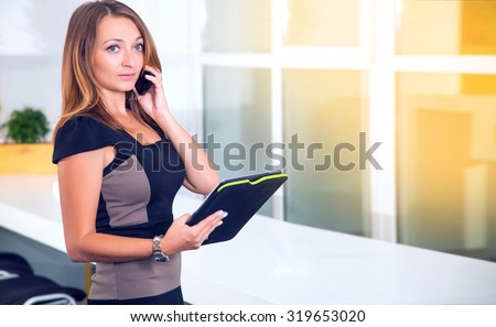 Young business woman talking on her phone while standing in front of an office room and windows. Businesswoman holding tablet pc computer and calling on cell phone. Communication technology.