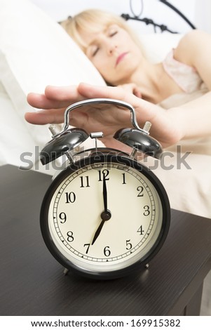A middle age woman reached for alarm clock in foreground which reads seven o\'clock.