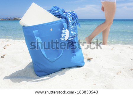 Beach bag on the sand, with book and foulard coming out from it and hair claw hanged on its handle, over walking girl and amazing clear water seascape