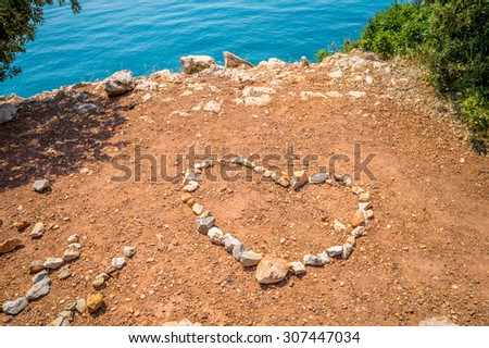 Heart made of stones on the red earth ground on the rocky beach, by the crystal clear blue sea, with some trees and bushes around it, with copy space for text