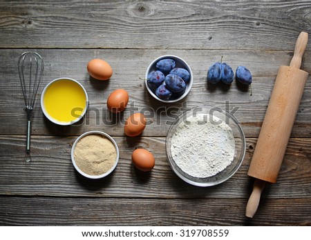 Baking cake in rural kitchen - dough recipe ingredients (eggs, flour, butter, sugar) and rolling pin  and plums on vintage wood table from above.