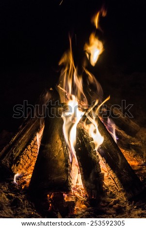 Camp fire burning, with the wooden logs in a typical triangle tee pee position, and flames reaching up into the black sky.