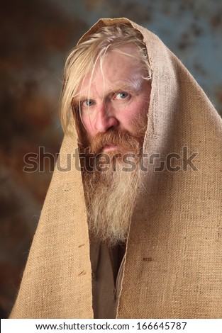 Portrait old beggar with a beard and mustache, wrapped in burlap, with pain and despair in his eyes, the studio on a brown-blue background