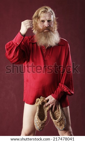 portrait of a man in a red shirt with long hair beard and mustache with a baton and bast shoes in hands studio on a burgundy background