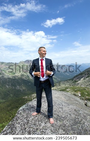portrait of a young man in a business suit at the top of the mountain on the background of mountains and blue sky on a sunny summer day