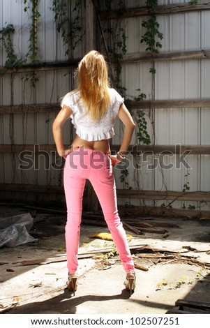 A lovely strawberry-blonde woman stands facing away from the camera wearing pink jeans in a long-abandoned warehouse facility.