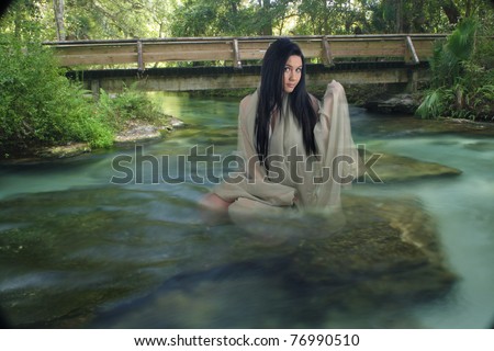 A lovely young teenage girl sits on a submerged rock, draped in sheer fabric, in a creek with crystal-clear flowing water.