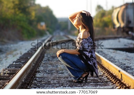 A lovely teenage Latina squats down on a railroad track, facing the sun with her eyes closed.