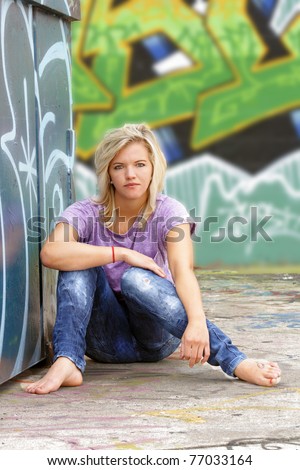 A lovely young blonde leans against a graffiti-covered garbage dumpster with a graffiti-covered wall behind her.
