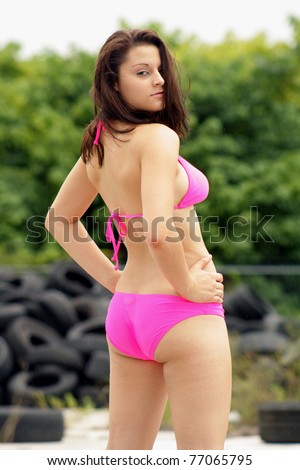 A beautiful rear view of a lovely young brunette wearing a hot-pink bikini outdoors with old tires in the background behind her.