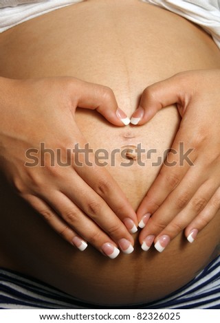 A woman who is eight months pregnant forms a heart over her belly with her hands.