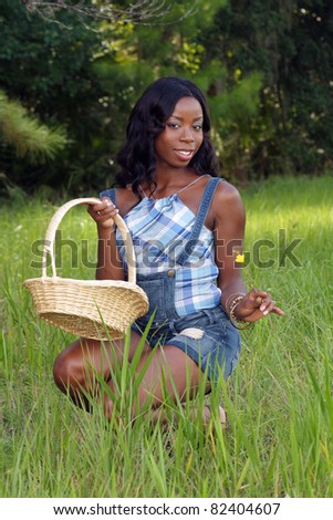 A lovely young black woman kneels in a grassy field, holding a woven basket.