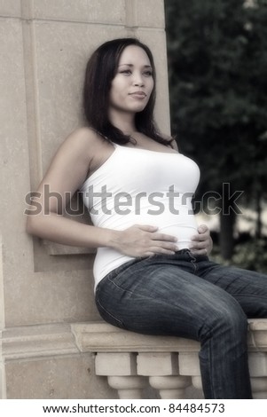 A lovely young multiracial woman five months pregnant, sits on an outdoor marble railing.  Desaturated with a light soft-focus effect.