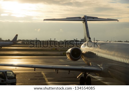 parked airplanes: large commercial plane is parked by the airport terminal (fuselage, turbine, wing and rear stabilizers visible), against the late afternoon sun and another airplane in the background