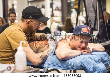 COPENHAGEN, DENMARK - FEBRUARY 8, 2014: a focused tattoo artist tattoos the back of a young man, whose face twists in pain, in a public location