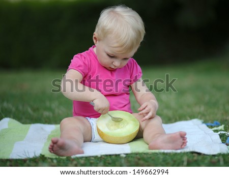 Cute baby girl sitting outdoors on a carpet eating melon with a spoon