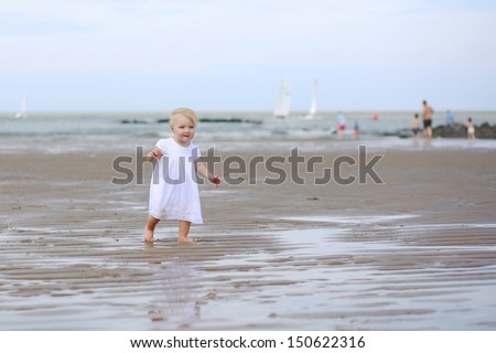 Happy baby girl in white dress walking on wet sand along coastline during tide of the North Sea, clear blue sky and yacht boats in the background, photo taken in Knokke, Belgium