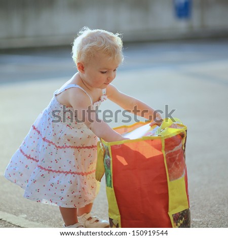 Cute little baby girl in beautiful dress looking inside big shopping back full of food products, fruits and vegetables. She is standing at a parking lot outside of supermarket store
