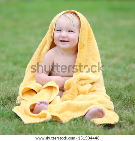 Lovely smiling baby having fun sitting on green grass in a garden wrapped in warm yellow towel drying after bathing in outdoors swimming pool