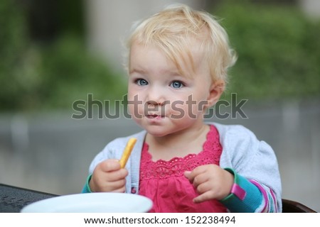 Cute little baby girl sitting on terrace at cafe eating delicious french fries