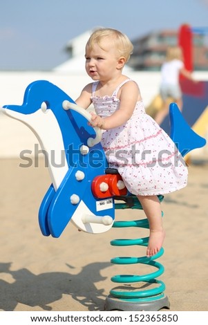Lovely happy blond baby girl plays outdoor at the beach sandy playground rocking on a spring blue dolphin