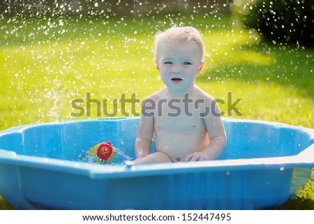 Cute funny little baby sitting in big blue plastic bath outdoors in a garden shade, splashing the water and playing with toys