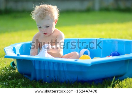 Funny lovely baby girl doing laundry playing with clothes sitting in a big blue plastic bath outdoors in a garden
