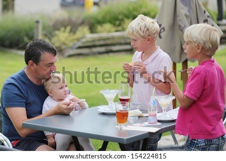 A family, father with two teenager sons and cute baby daughter are relaxing together outdoors in cafe on summer terrace eating ice cream and drinking beer and ice tea