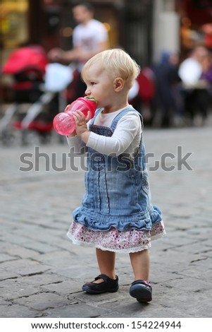 Funny little baby girl walks on a busy street drinking water from plastic feeding bottle, in background people are sitting eating in cafe
