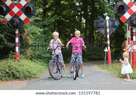 Two boys cycling on bikes at training playground in park, they learn how to safely cycle in city by looking at educational traffic signs and stopping at red light; baby sister plays role of pedestrian