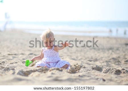 Sweet blond little baby girl in beautiful white dress plays with water and sand sitting at a shore of the sea on a long calm empty peaceful beach on a warm sunny summer day