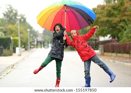 Two children, teenager twin brothers having fun outside jumping on the street under rain sharing together colorful umbrella