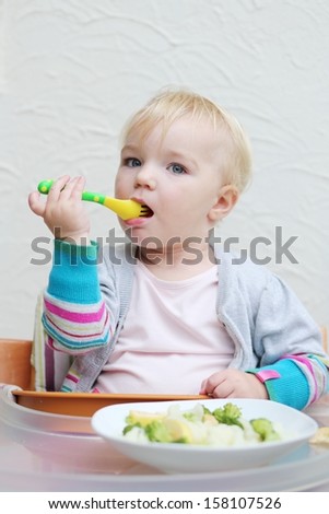 Lovely little baby girl eating fresh fruits and vegetable holding plastic fork sitting in high feeding chair