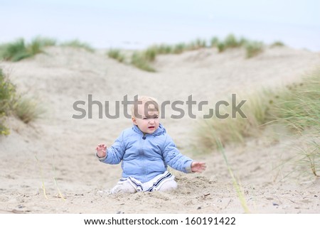 Active little baby girl in warm coat plays with sand in the dunes