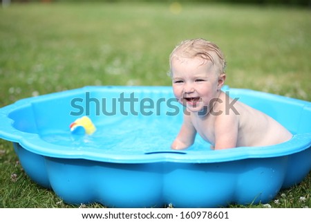 Cute happy baby laughing sitting inside of little plastic bath in the garden on a hot summer day