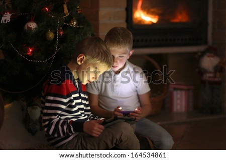 Two twin brothers sitting at the fireplace on a cozy winter evening nearby the christmas tree choosing gifts from online catalog or playing with tablet pc and smart phone