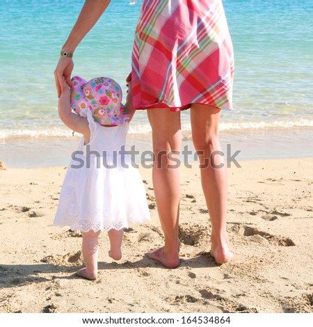 Mother and baby: young woman is teaching her sweet toddler daughter to walk her first steps on a sandy tropical beach