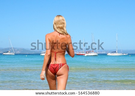 Beautiful young blonde slim woman walking into the sea, looking at white yacht boats at the horizon