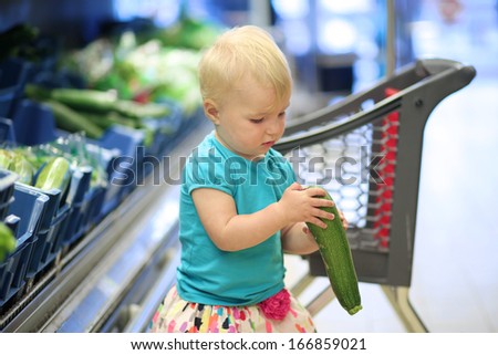 Cute blonde toddler girl looking at zucchini picked up from a shelf in vegetables department in a supermarket