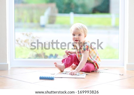Cute little blonde toddler girl playing with puzzles sitting on the tiles floor next to a big window