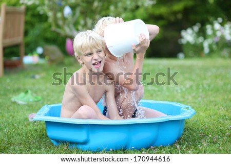 Two cute boys, twin brothers, playing with bucket of water in little plastic bath outdoors in the garden at the backyard of the house