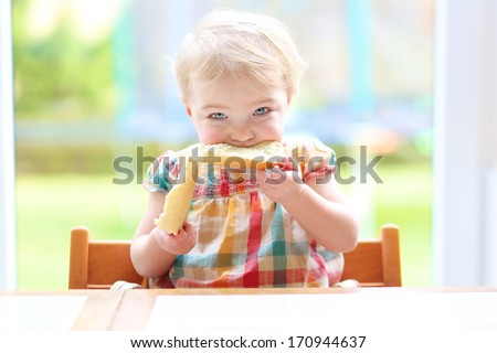 Funny blonde toddler girl eating tasty bread with butter sitting in the kitchen nearby big window with garden view