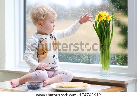 Cute blonde toddler girl enjoying pancakes sitting on the kitchen table next to a big window with narcissus flowers on a rainy spring day